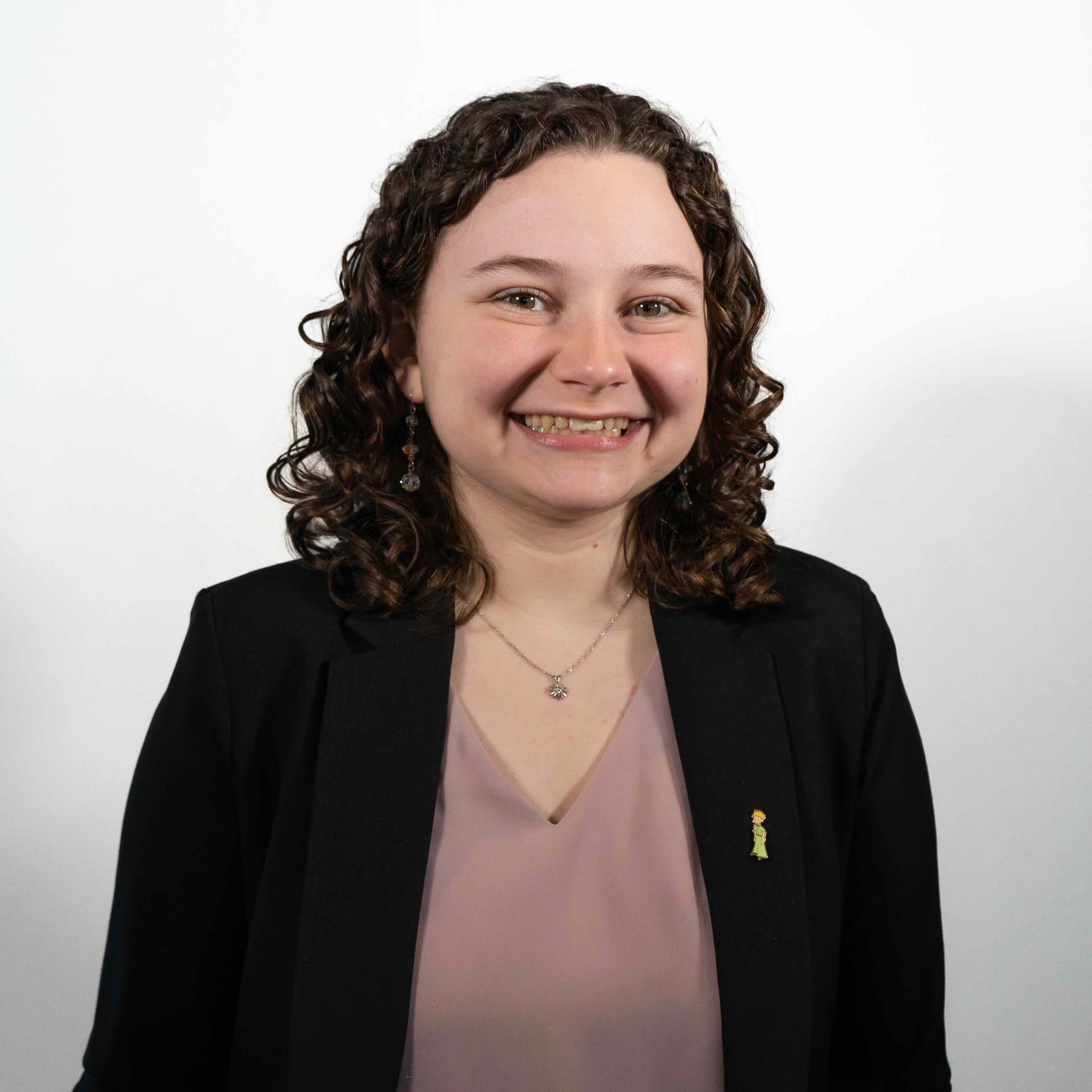 Smiling girl with curly hair, dressed professionally with an enamel pin of The Little Prince on her suit jacket.